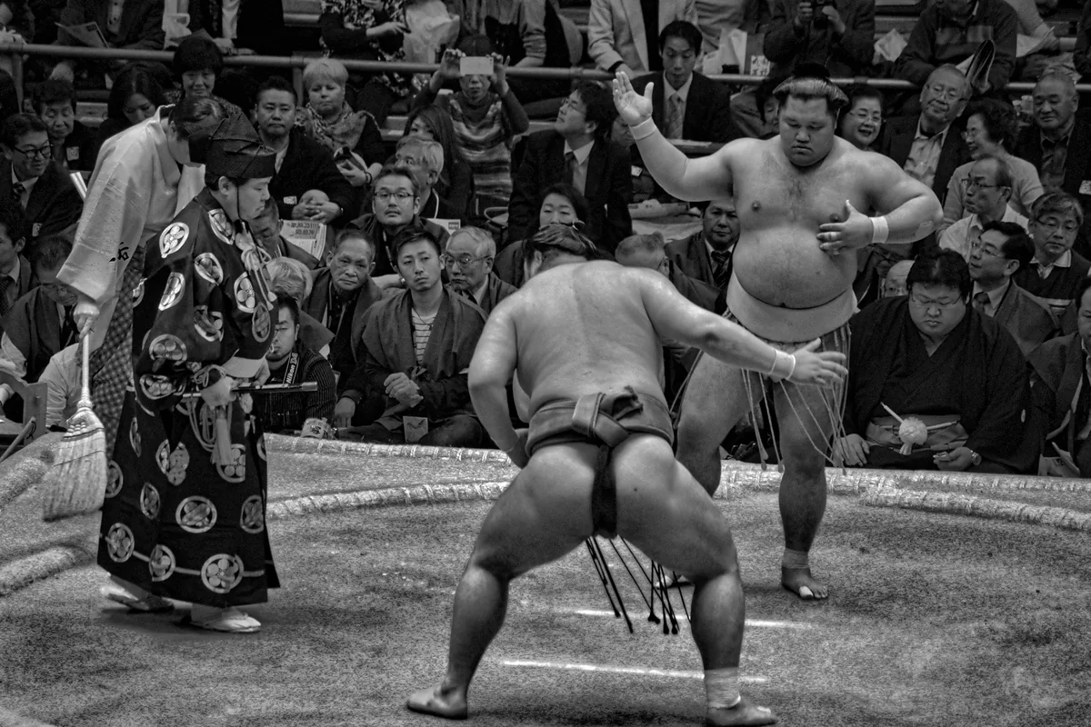 A sumo wrestler mid-bout in front of a seated crowd, with a referee in ceremonial dress nearby, photographed in Osaka, Japan