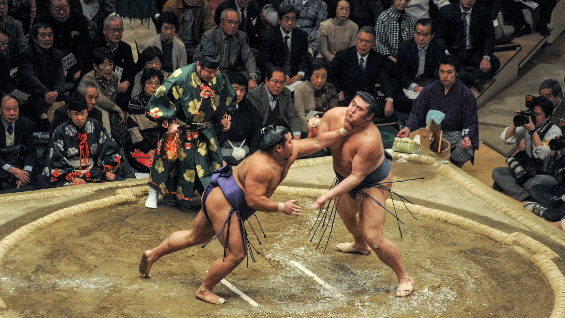 Two sumo wrestlers grappling on a clay ring in front of a seated crowd, with a referee in ceremonial dress looking on